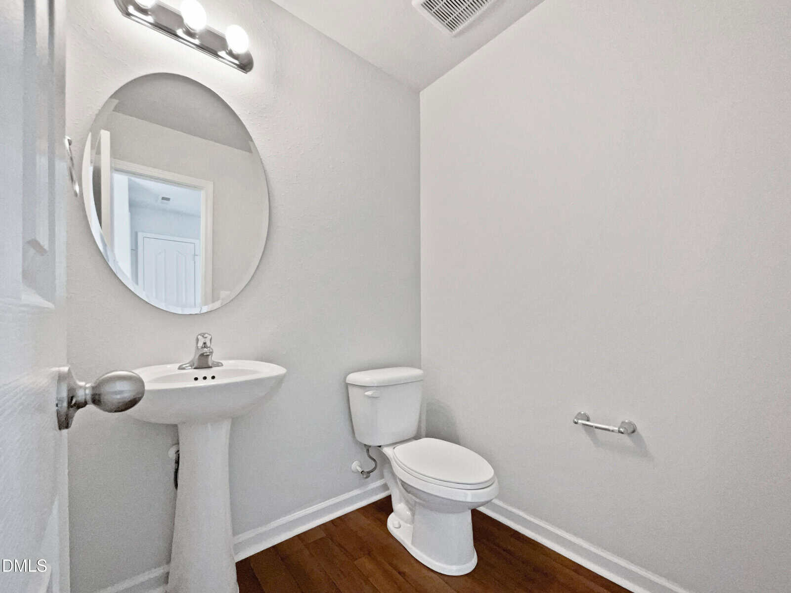 A small bathroom with white walls, a pedestal sink, a round mirror above it, a toilet, a wall-mounted light fixture, a toilet paper holder, and wood flooring.