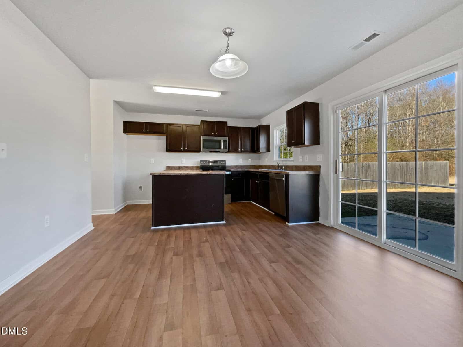 A modern kitchen with dark cabinets, stainless steel appliances, and an island, set in an open space with wood flooring. Large sliding glass doors lead to a backyard with a wooden fence and trees.
