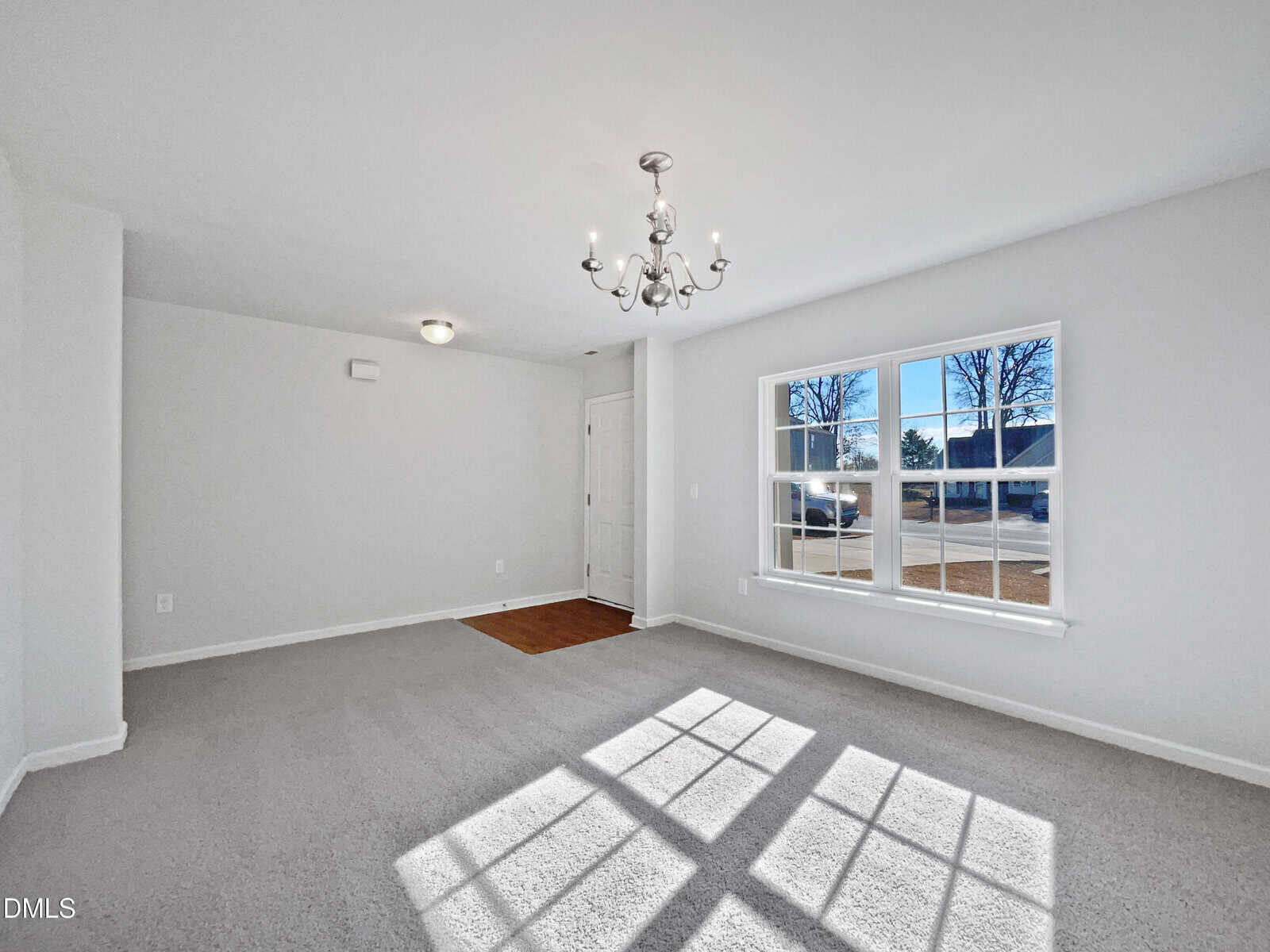 A bright, empty room with light gray carpet, white walls, a chandelier, a large window letting in sunlight, and a door with a small entry area featuring wood flooring.