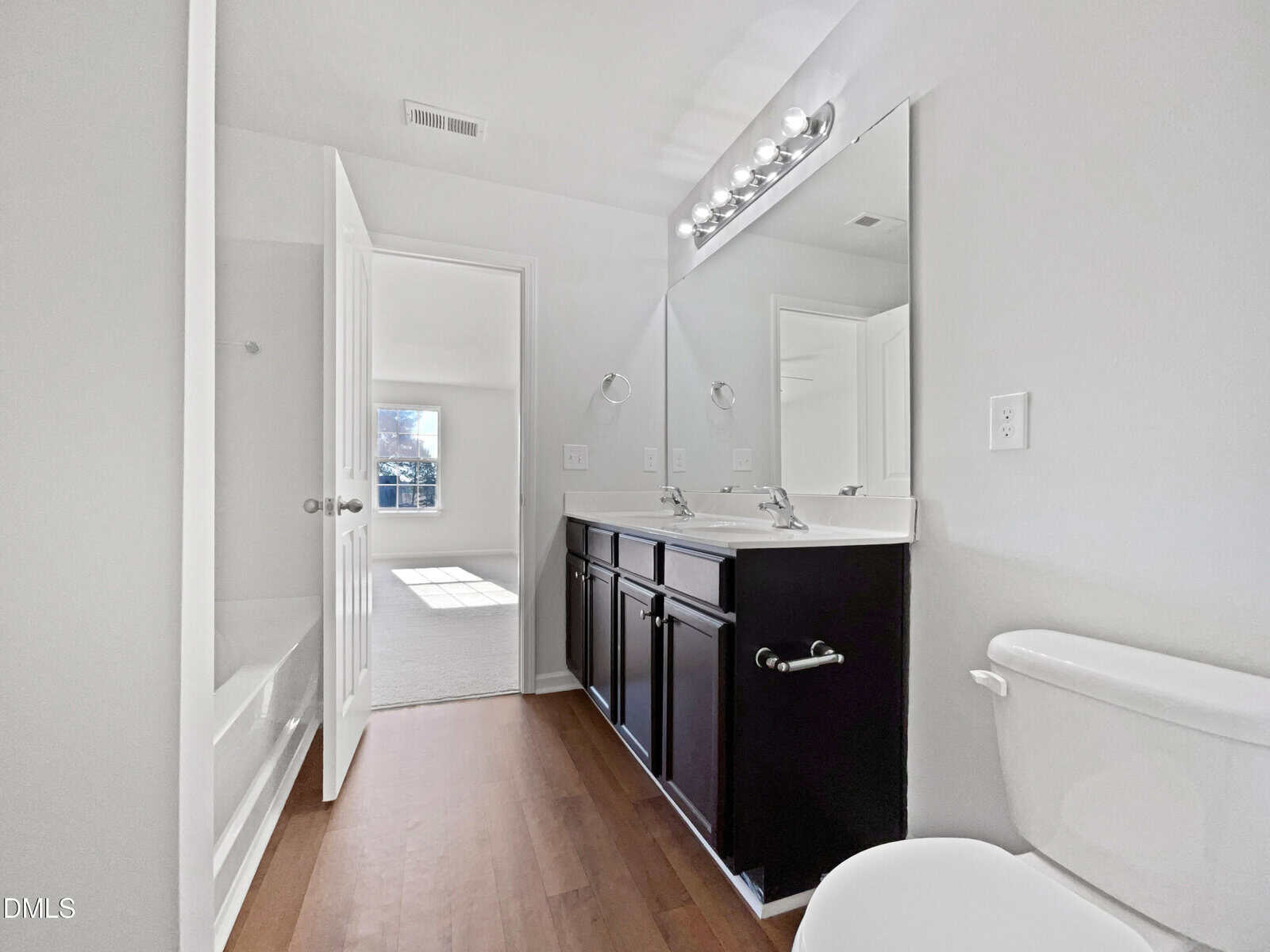 A modern bathroom with a double sink vanity, large mirror, bathtub with shower, and toilet. The floor is wood, and a door leads to a bright carpeted bedroom with sunlight coming through a window.