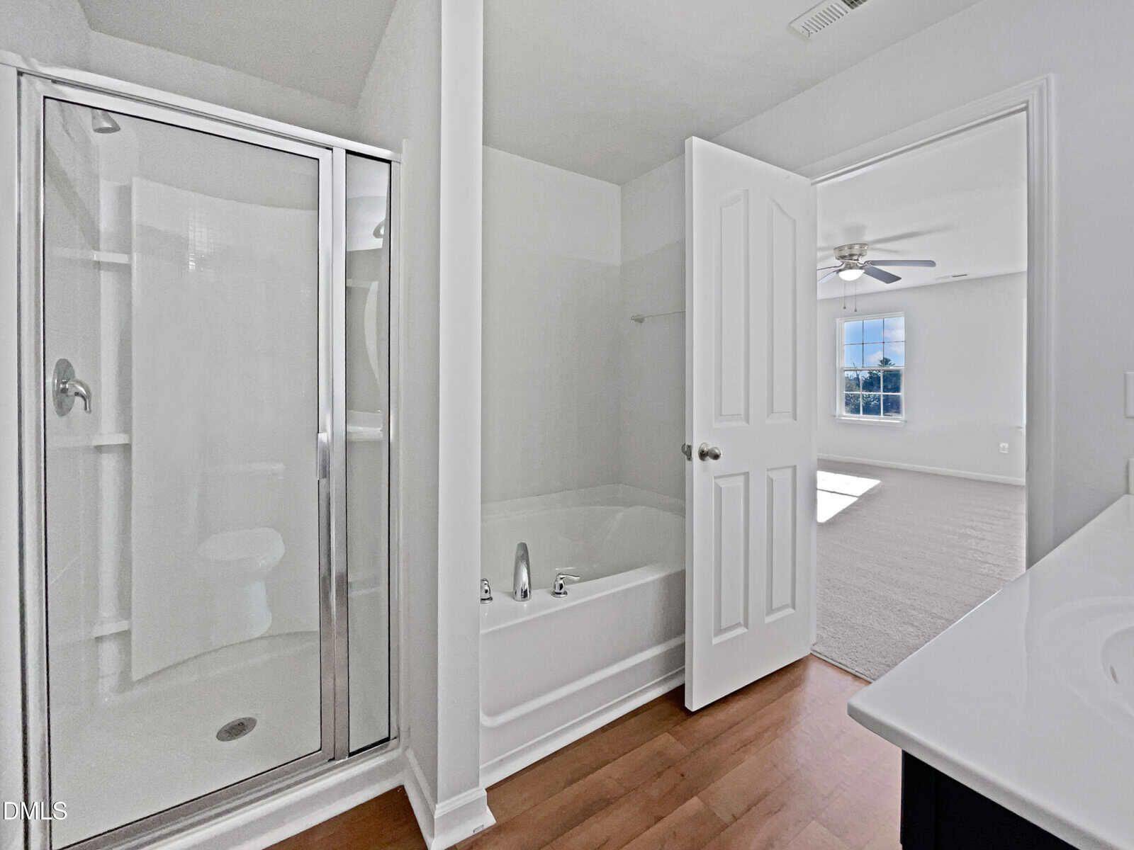 White bathroom with a glass shower, bathtub, sink with vanity, and an open door leading to a bright, carpeted bedroom with a ceiling fan and window.