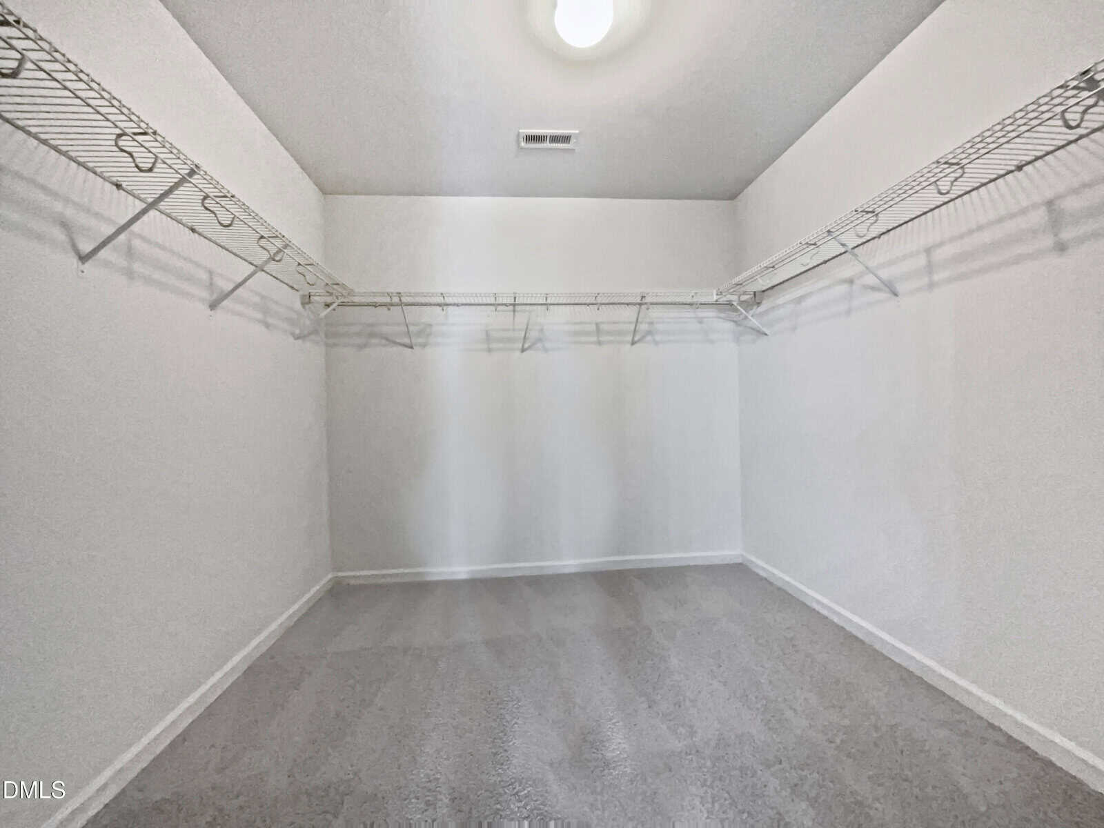Empty walk-in closet with white walls, beige carpet, ceiling light, and wire shelves lining two walls. The space is clean, well-lit, and spacious.