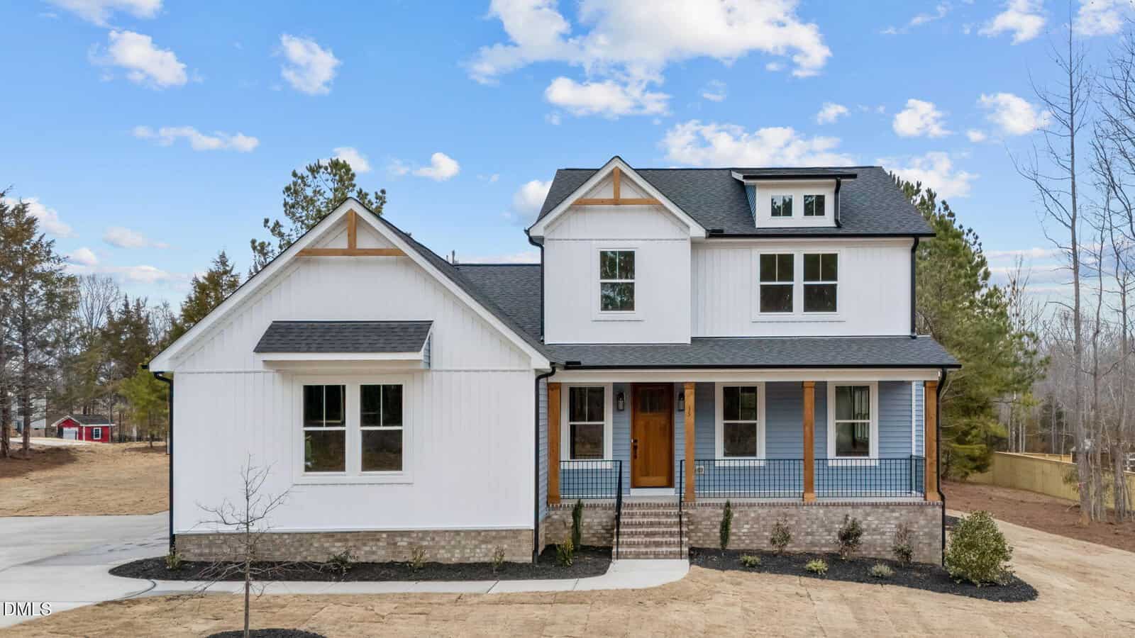A two-story modern farmhouse with white siding, black roof, and wooden accents. The house has a front porch with columns, brick steps, and a landscaped yard with young plants and trees. The sky is clear with scattered clouds.