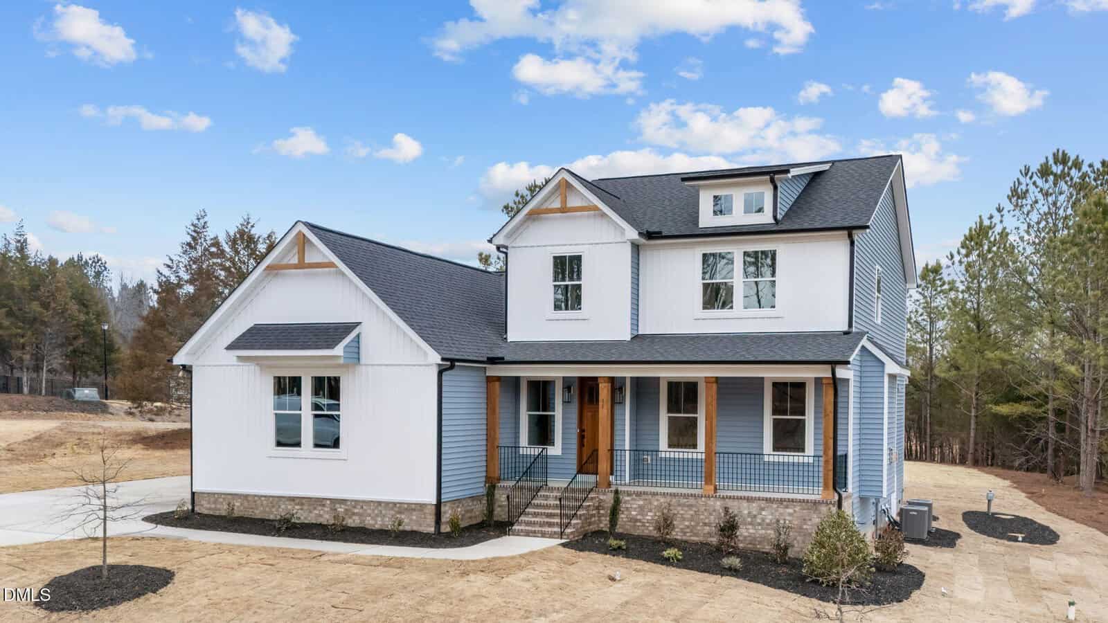 A two-story modern farmhouse with blue and white siding, a covered front porch with wooden posts, brick foundation, and a neatly landscaped yard, set against a backdrop of trees and a partly cloudy sky.