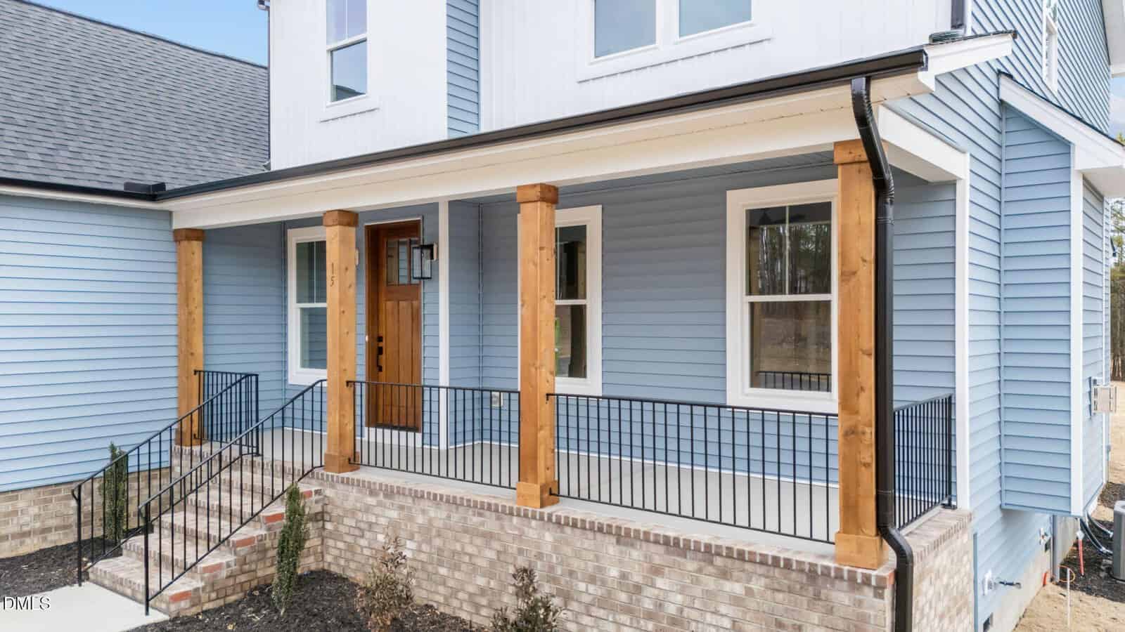 A modern house exterior with blue siding, white trim, and a wooden front door. The porch features wooden columns, black metal railings, brick foundation, and steps leading up to the entrance.