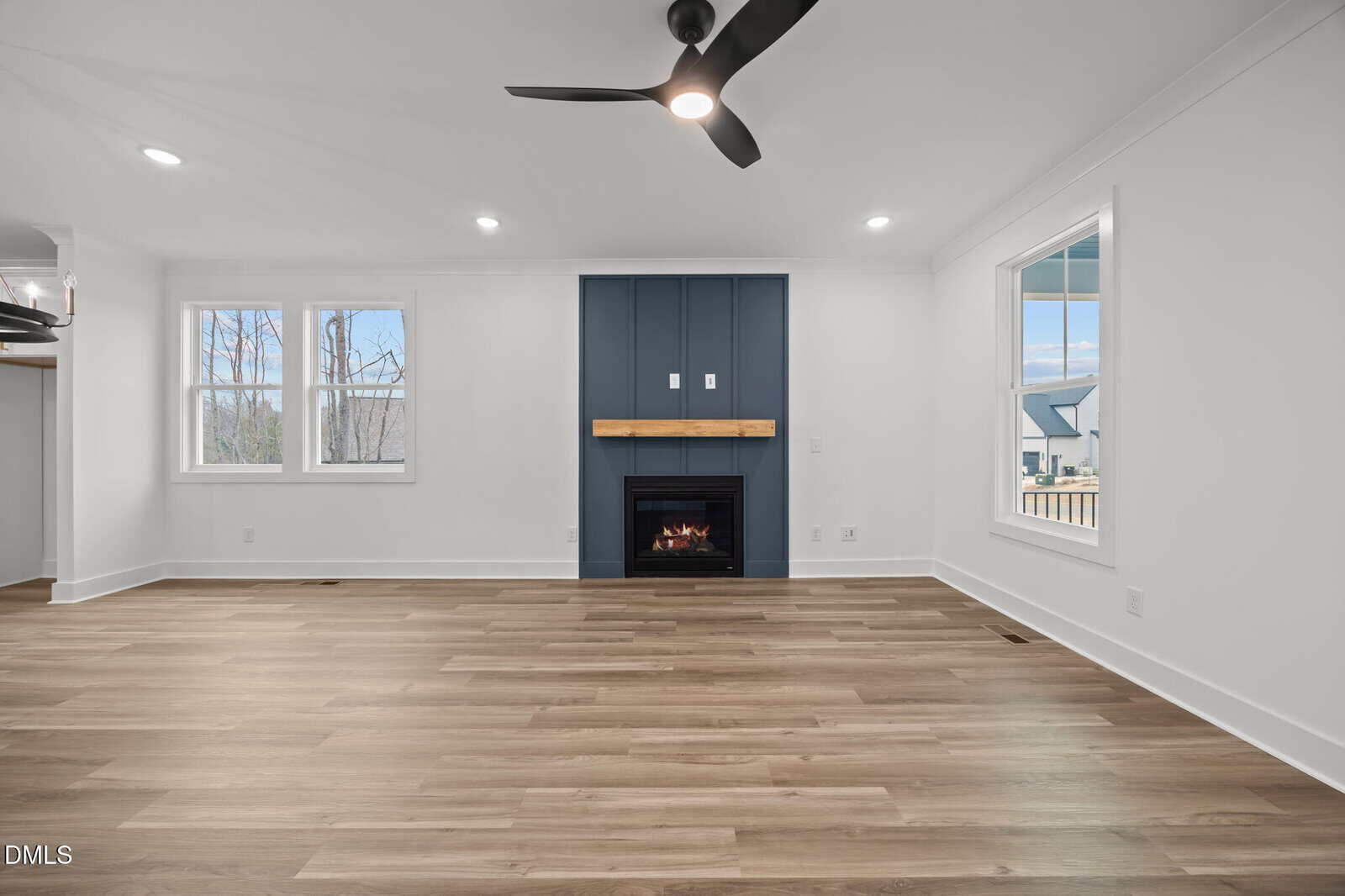 A modern living room with light wood floors, white walls, a black ceiling fan, large windows, and a dark blue accent wall with a fireplace and wooden mantel.