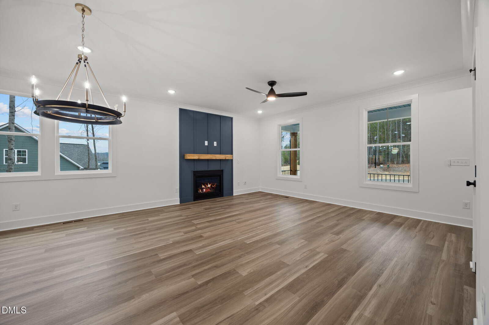 Modern empty living room with wood floors, white walls, large windows, a ceiling fan, chandelier, and a fireplace with a blue accent wall and wood mantel.