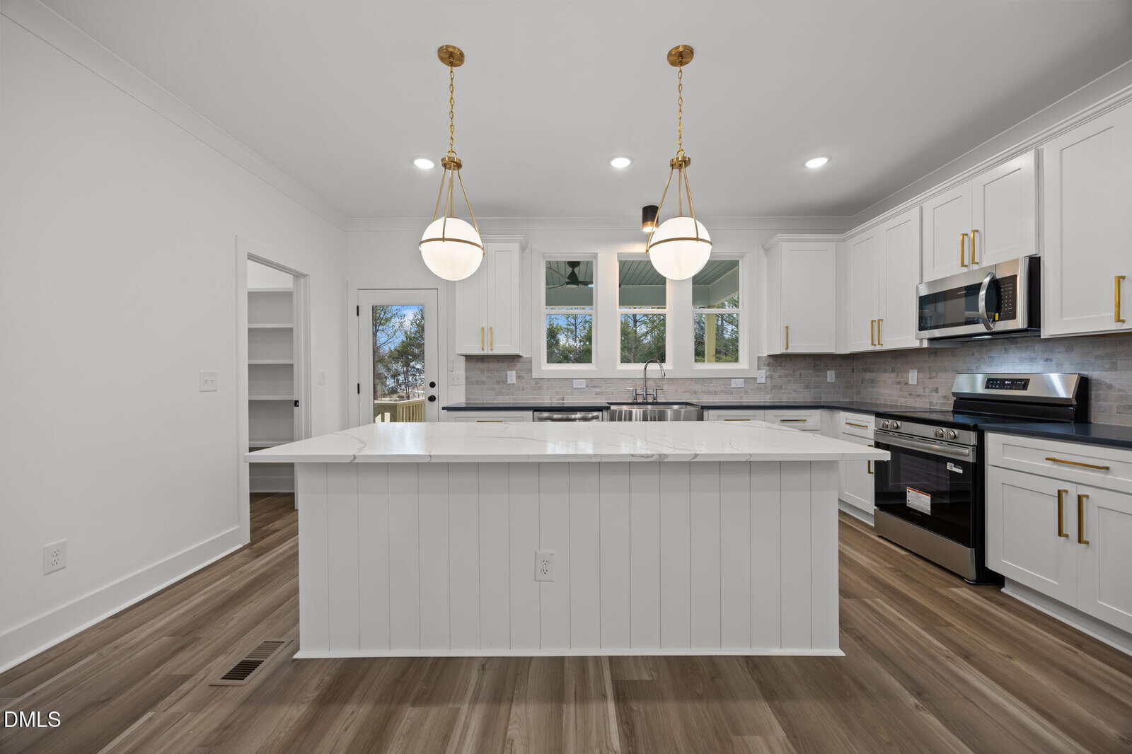 A modern kitchen with white cabinets, a large white island with a marble countertop, stainless steel appliances, wood flooring, pendant lights, and three windows above the sink.
