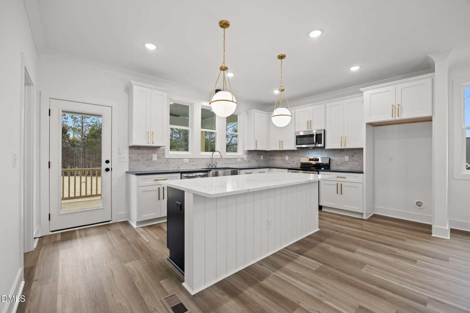 A modern kitchen with white cabinets, a central island, stainless steel appliances, pendant lights, light wood flooring, and large windows. A door on the left leads outside, and there is an empty space for a refrigerator.