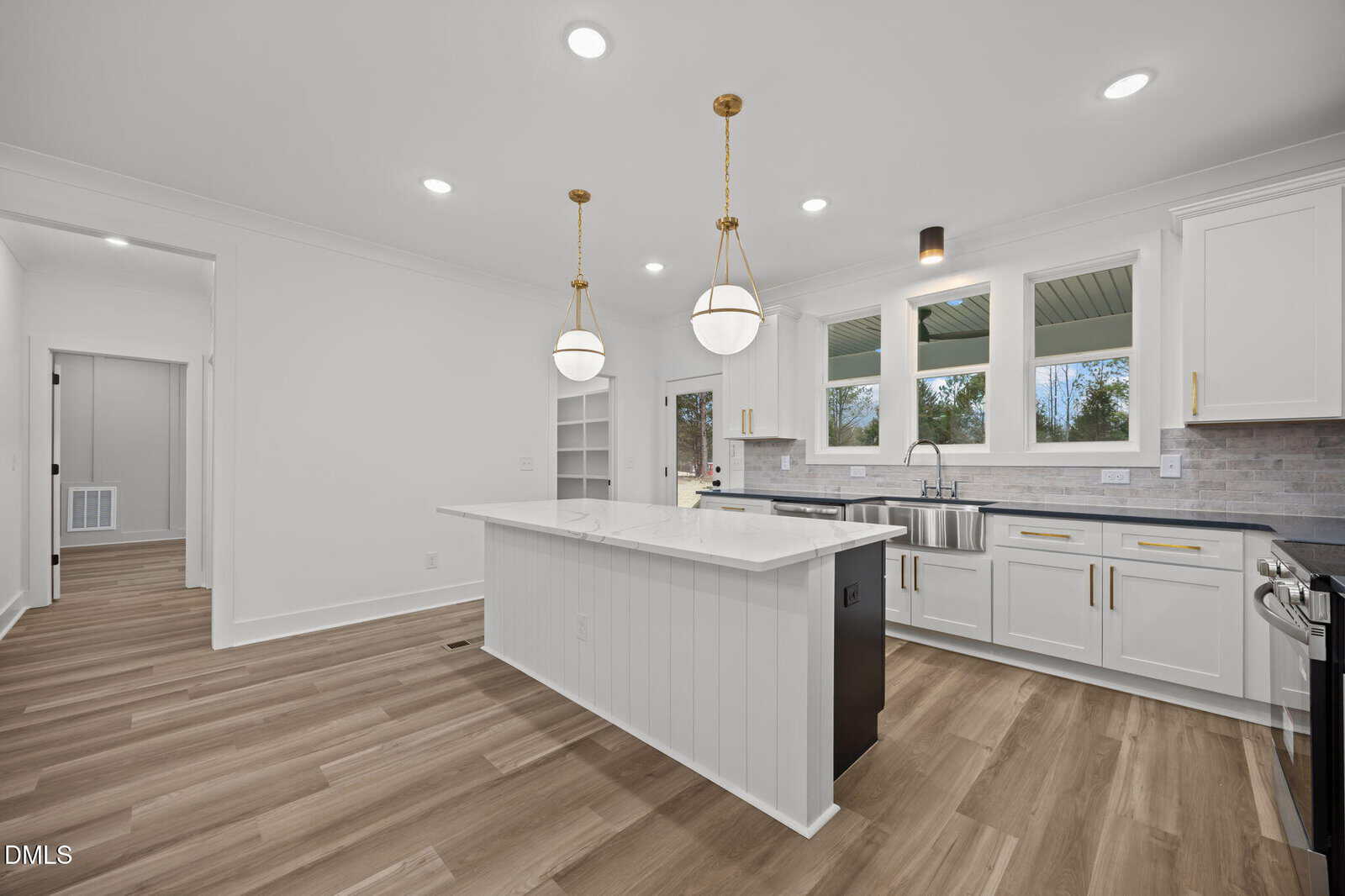 Modern kitchen with white cabinets, a large island with a sink, stainless steel appliances, two pendant lights above the island, light wood flooring, and windows above the farmhouse-style sink.