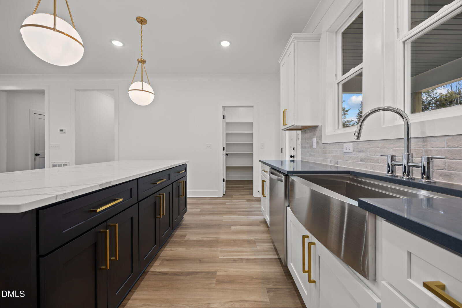 Modern kitchen with a large island, black lower cabinets with gold handles, white upper cabinets, farmhouse sink, two pendant lights, wood flooring, and a walk-in pantry visible in the background.