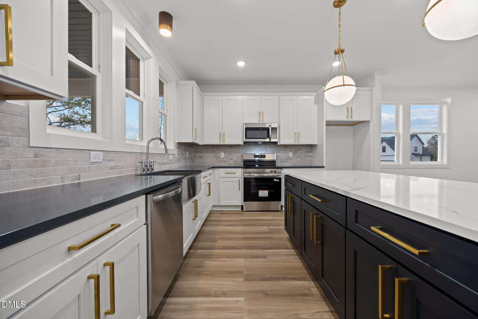 Modern kitchen with white upper cabinets, black lower cabinets, stainless steel appliances, light wood flooring, marble island countertop, brass hardware, and pendant lights, with windows providing natural light.