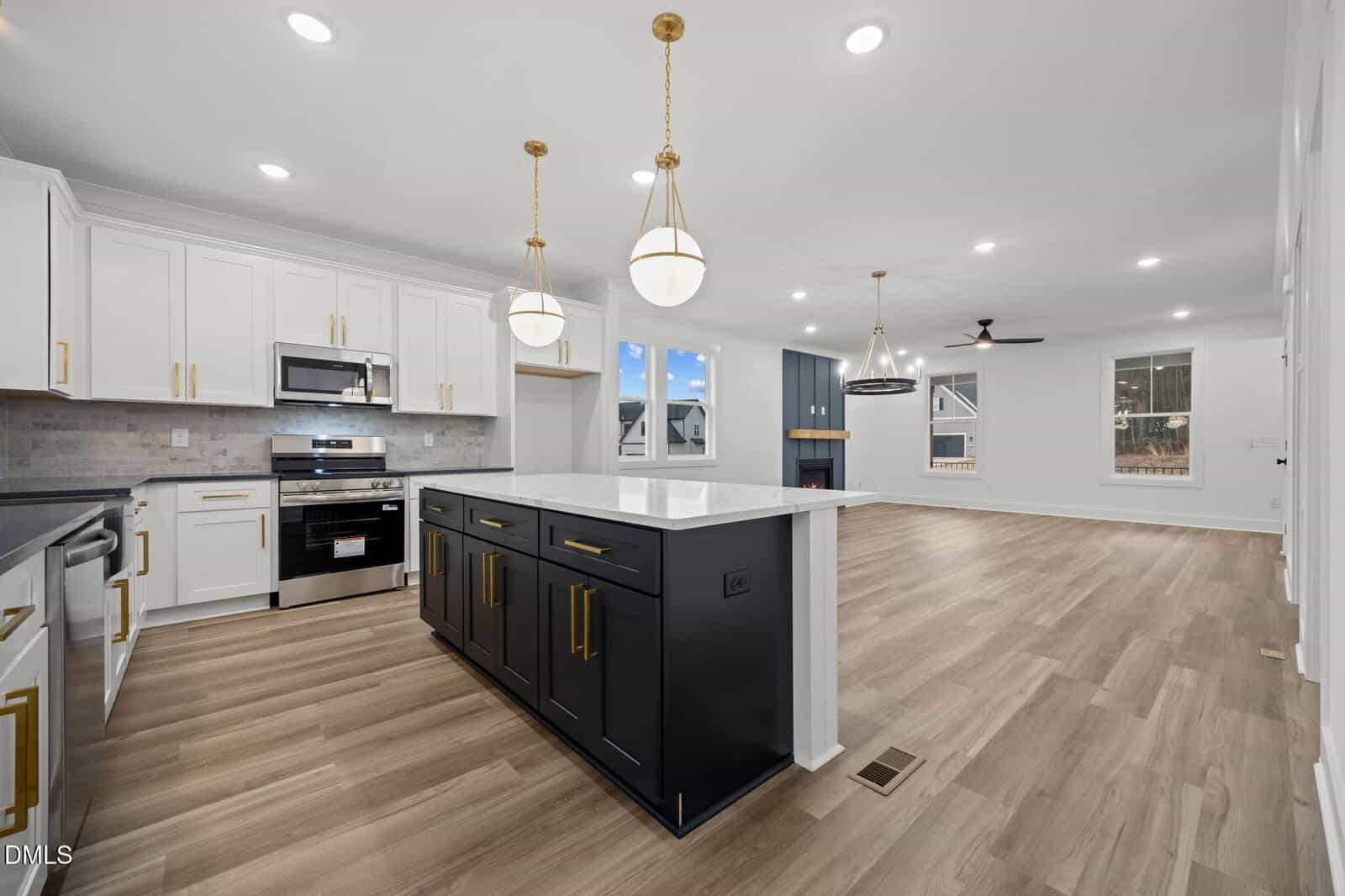 Modern open-concept kitchen with white cabinets, stainless steel appliances, black island with gold hardware, pendant lights, and light wood floors, opening to a living area with fireplace and large windows.