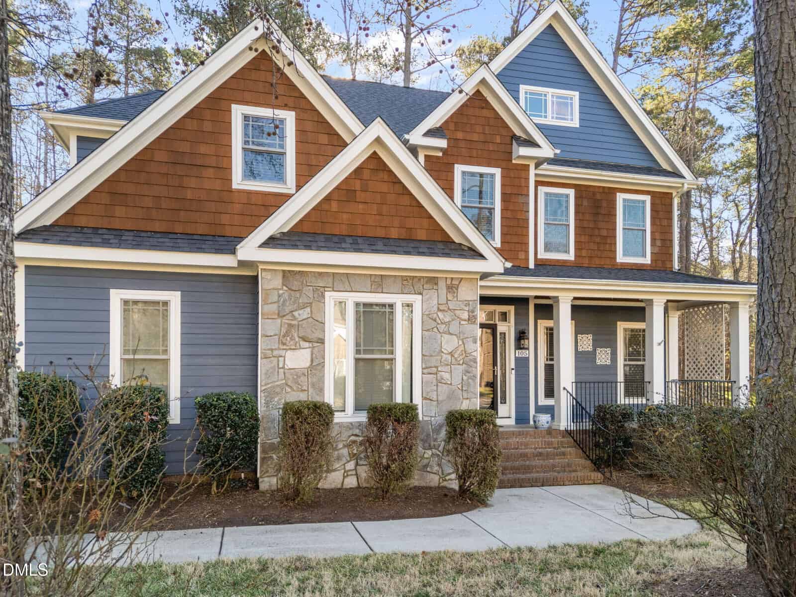 A two-story suburban house with brown shingle and blue siding, stone accent wall, white trim, multiple gables, large windows, a small porch with black railings, and trimmed bushes in the front yard.