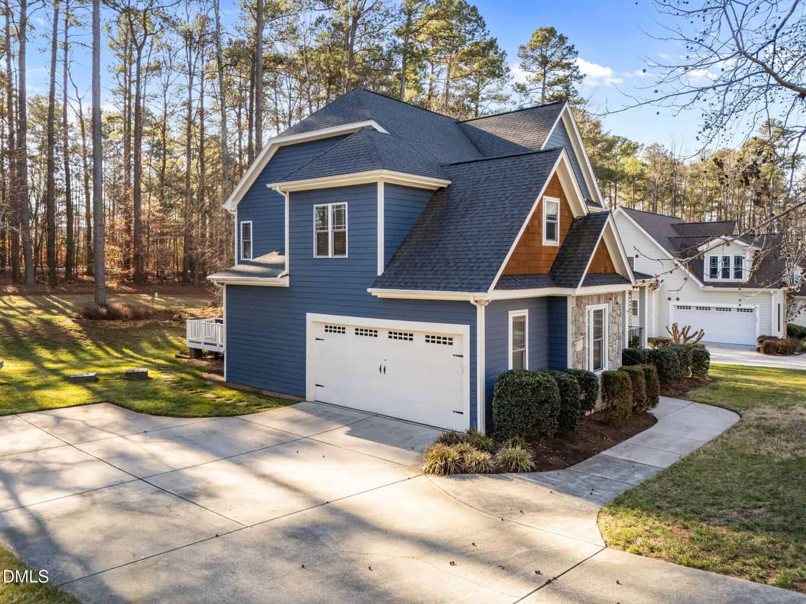 A two-story blue house with white trim and a double garage sits on a curved driveway, bordered by well-maintained grass and shrubs. Tall trees line the background under a clear sky.