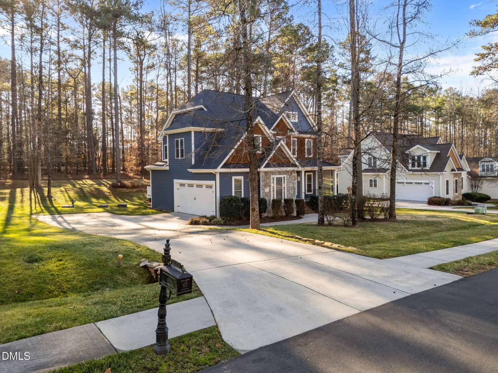 A two-story house with blue siding and brown accents sits next to a similar neighboring home. Both have attached garages and well-maintained lawns. Tall trees line the background and a black mailbox is by the curb.