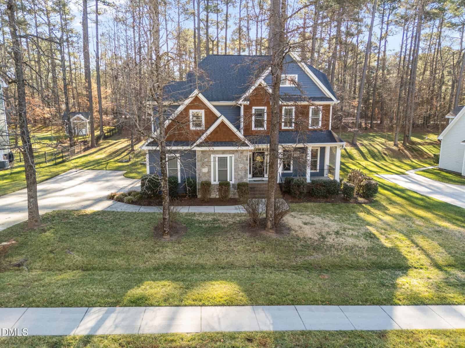 A two-story house with a front porch and gabled roof is surrounded by trees and a lawn. A driveway curves on both sides of the house, and a sidewalk runs along the street in front.