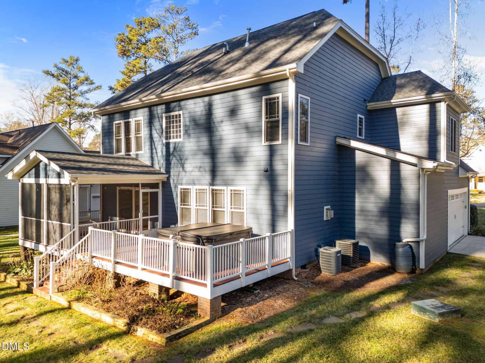 A two-story blue house with white trim features a screened porch, large raised deck with a hot tub, two air conditioning units, a rain barrel, and a two-car garage. The yard is grassy with some trees and landscaping.