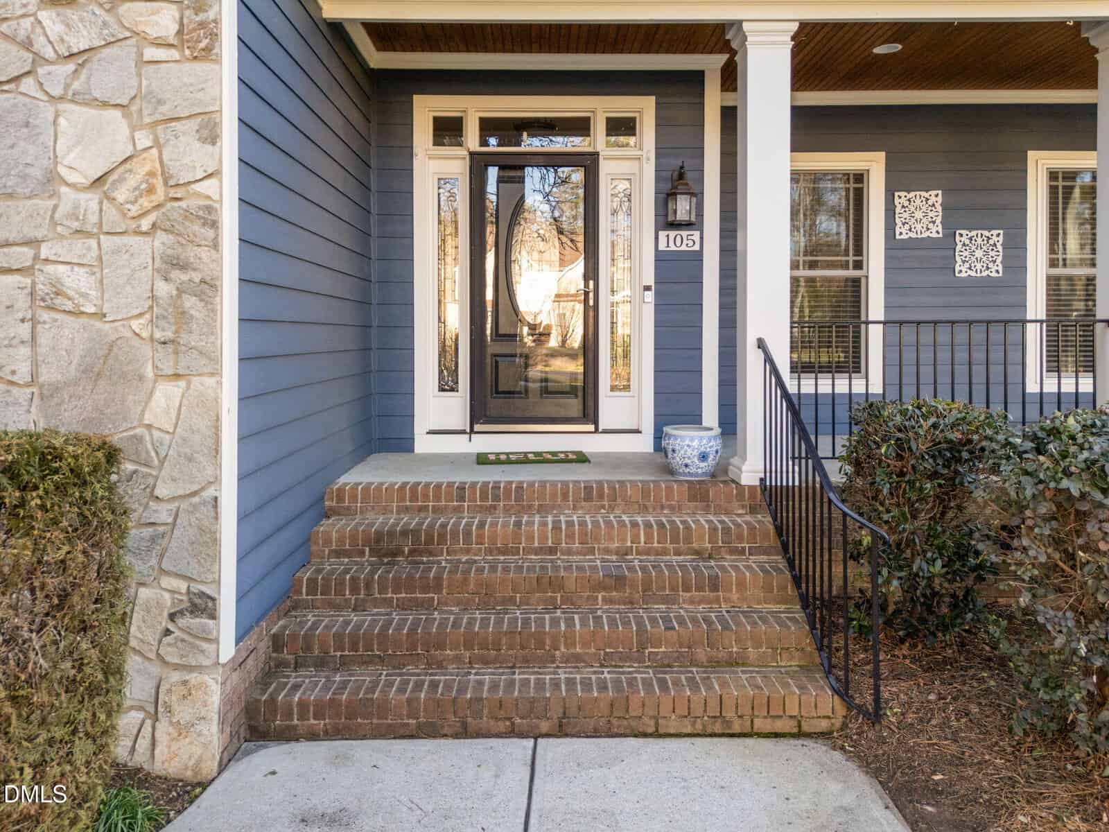 A brick staircase with six steps leads to a glass front door of a blue house with white trim. A black railing, potted plant, and house number 105 are visible. Shrubs and a stone wall frame the entry.