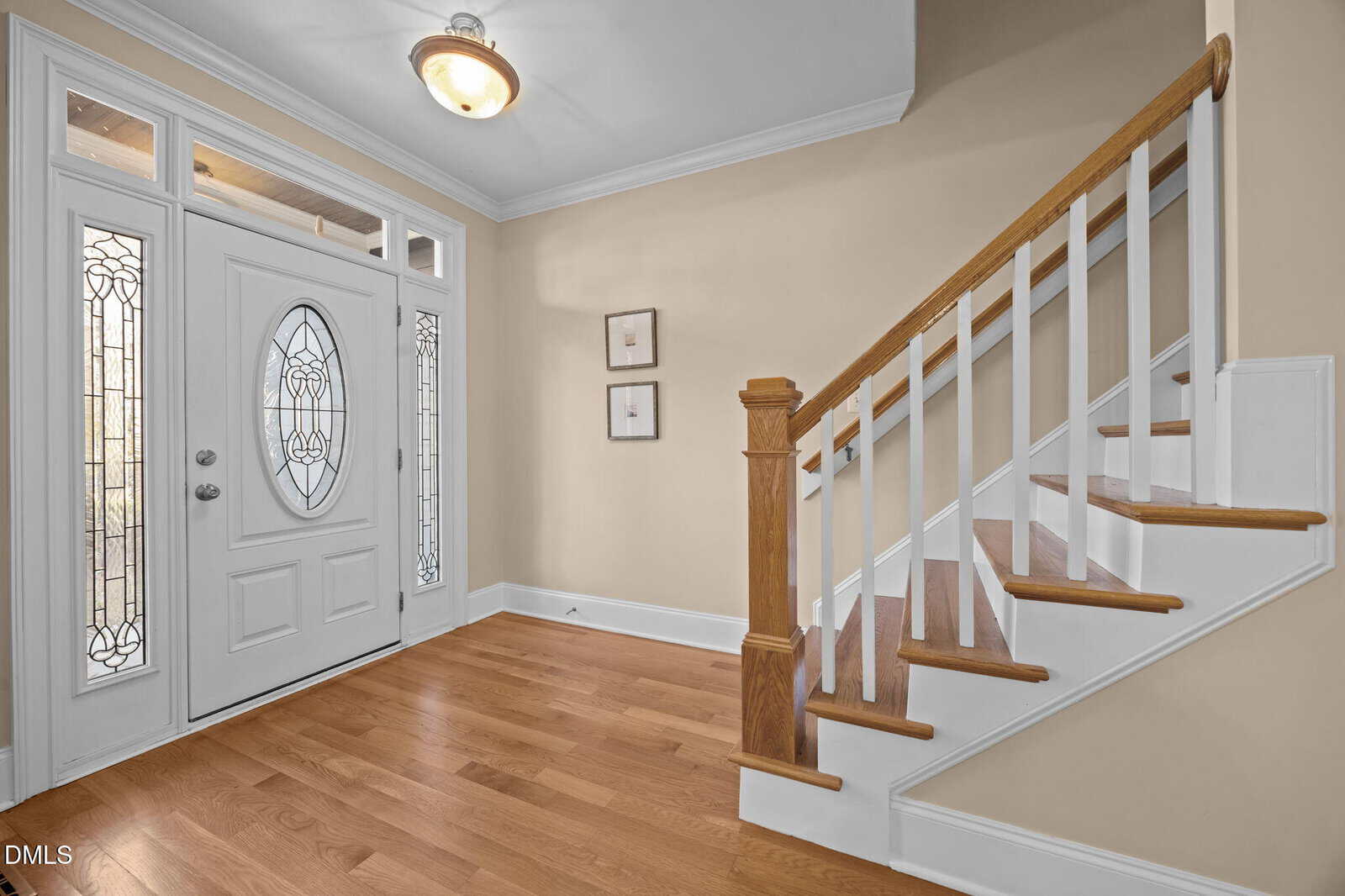 A bright foyer with hardwood floors, a white front door featuring decorative glass panels, two framed pictures on a beige wall, and a wooden staircase with white risers and balusters.