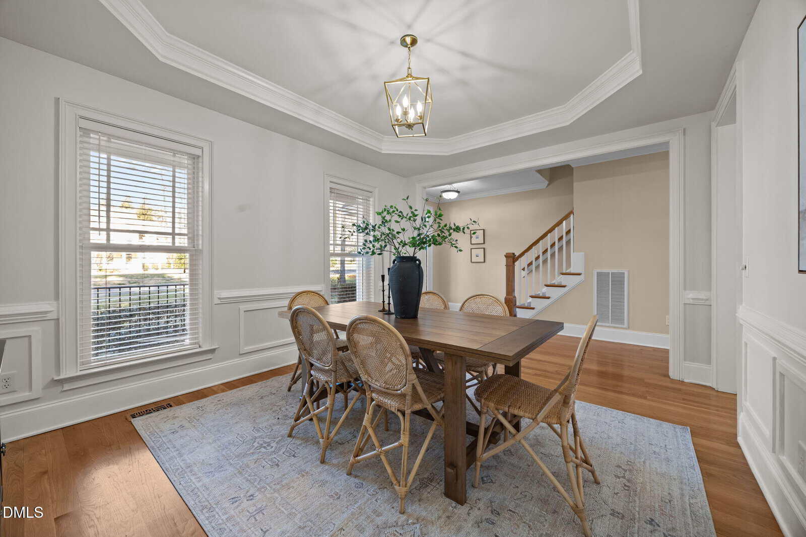 A dining room with a wooden table, six wicker chairs, a blue and white rug, a black vase with green branches, hardwood floors, white walls, large windows, and a gold chandelier. Stairs are visible in the background.