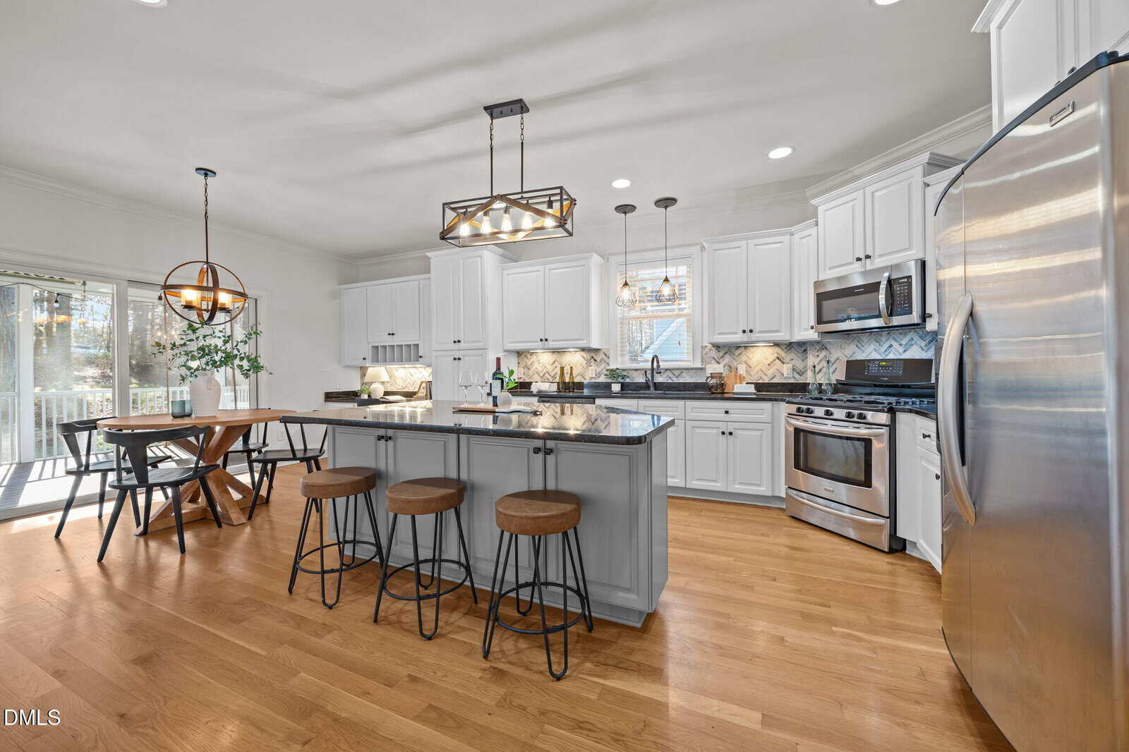 A modern kitchen with white cabinets, stainless steel appliances, a center island with three stools, a wooden dining table with chairs, and hardwood floors. Pendant lights hang over the island and table.