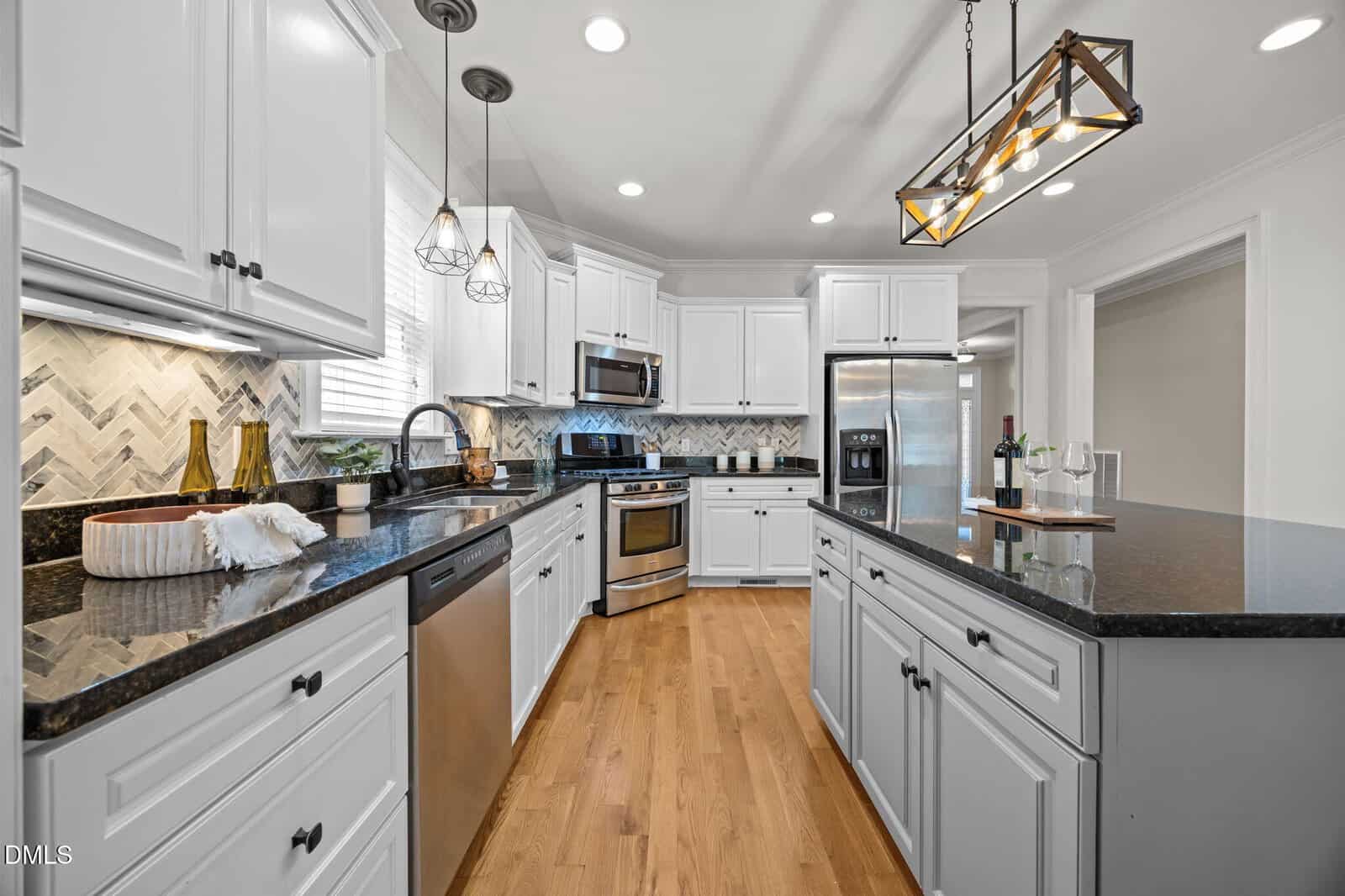 Modern kitchen with white cabinets, stainless steel appliances, black countertops, hardwood floor, an island with pendant lighting, and a herringbone tile backsplash. Wine and plants decorate the counters.
