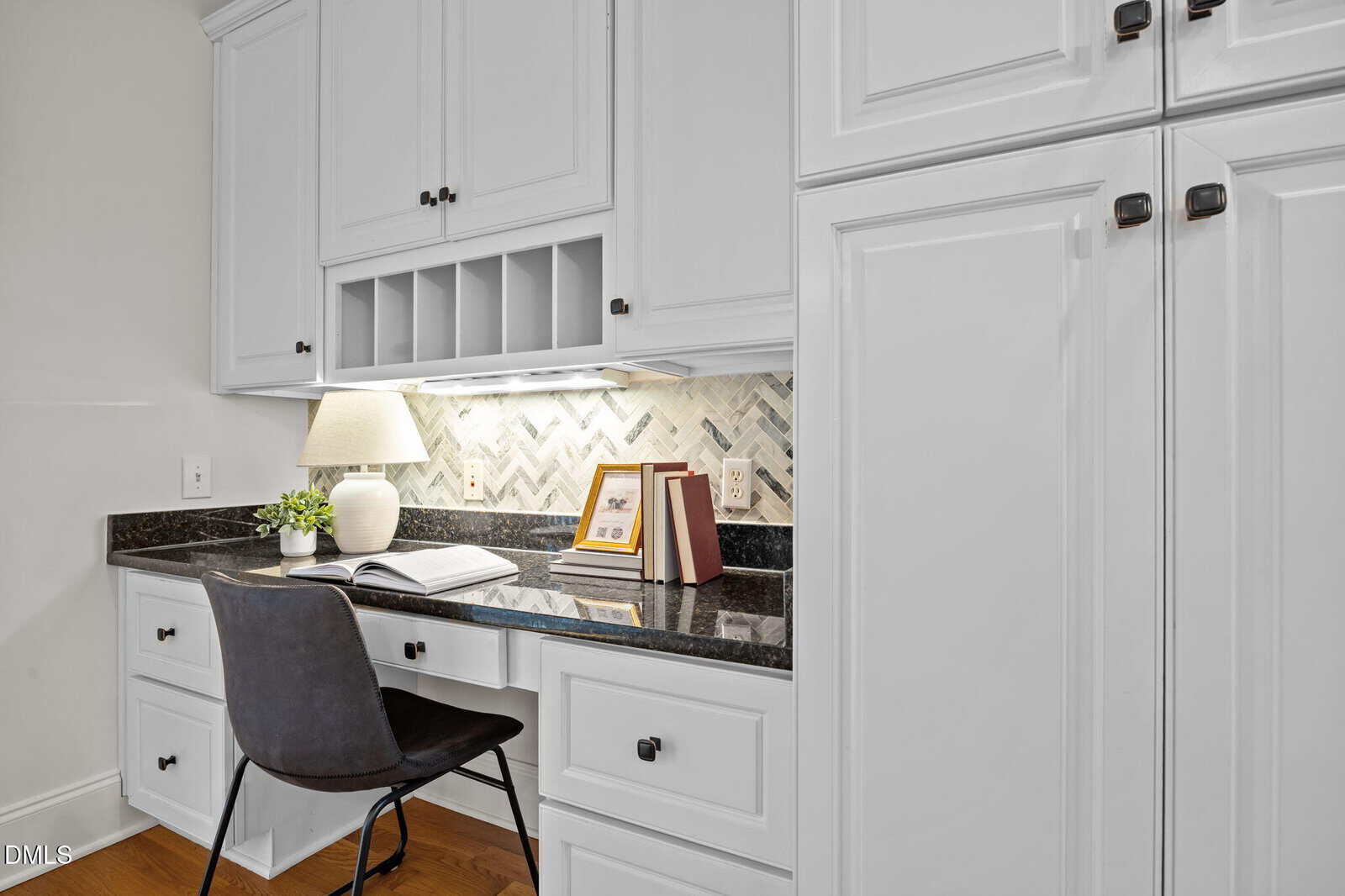 A built-in white desk with a black chair, open book, two closed books, lamp, and herringbone tile backsplash under cabinets. The desk sits against a wall with white cabinetry and dark countertop.