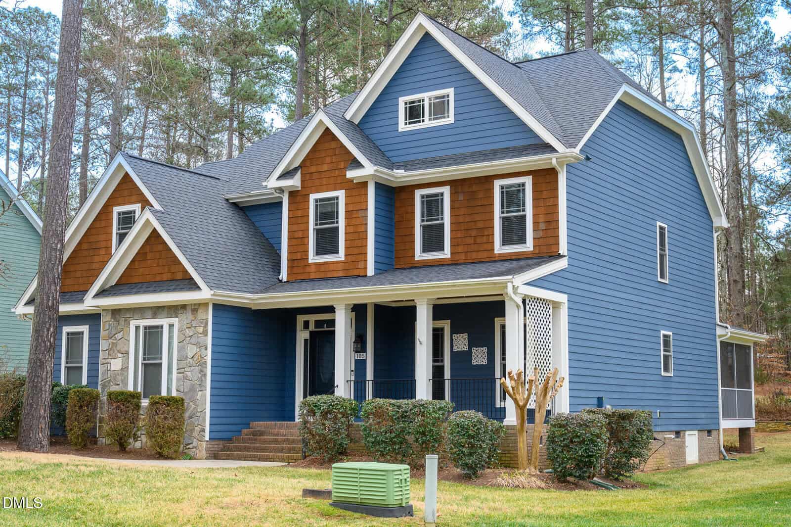 This two-story house at 105 Patterson Drive in Youngsville features blue siding, brown shingle accents, stonework on the lower front, a covered porch, gabled roofs, trimmed bushes, and is surrounded by tall trees and a lawn.