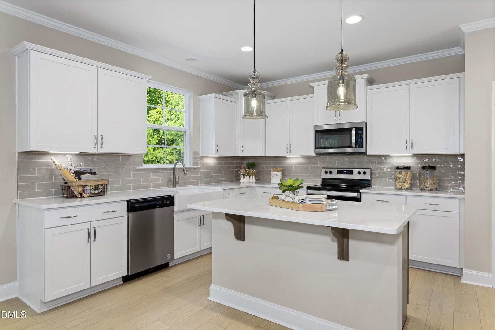 Modern kitchen with white cabinets, stainless steel appliances, light wood flooring, a large island with a white countertop, pendant lights, and a window letting in natural light above the sink.
