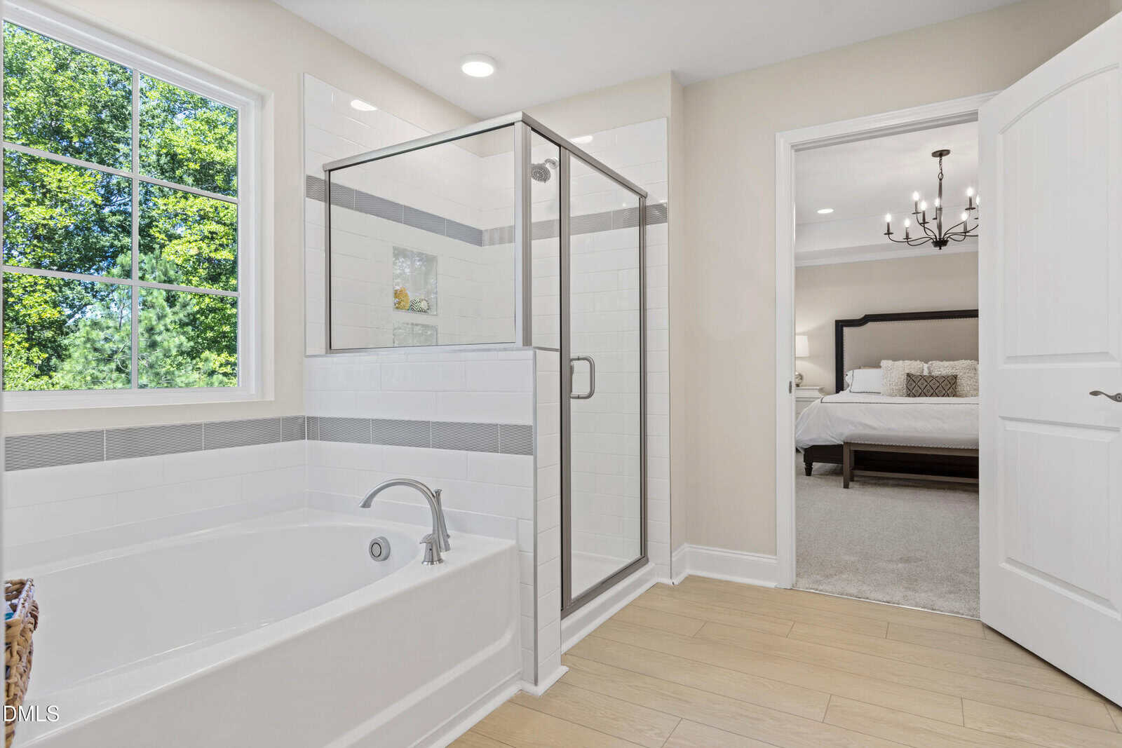 Modern bathroom with a glass shower, white bathtub, and light wood flooring. A window shows green trees outside. An open door leads to a bedroom with a chandelier and neatly made bed.