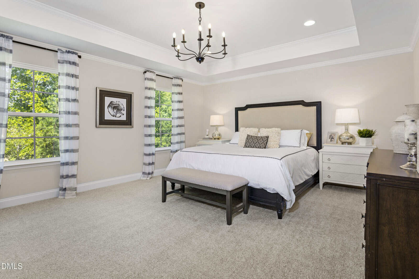 Spacious, bright bedroom with a large bed, two nightstands with lamps, a dark bench at the foot of the bed, two windows with striped curtains, a chandelier, and neutral decor.