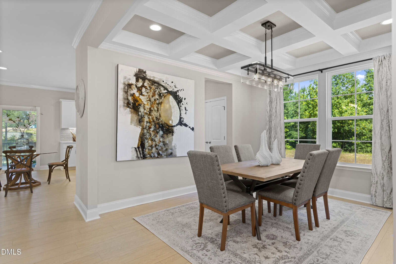 Modern dining room with a wooden table, six gray upholstered chairs, a large abstract painting on the wall, and a light fixture above. Large windows let in natural light, showing a green outdoor view.