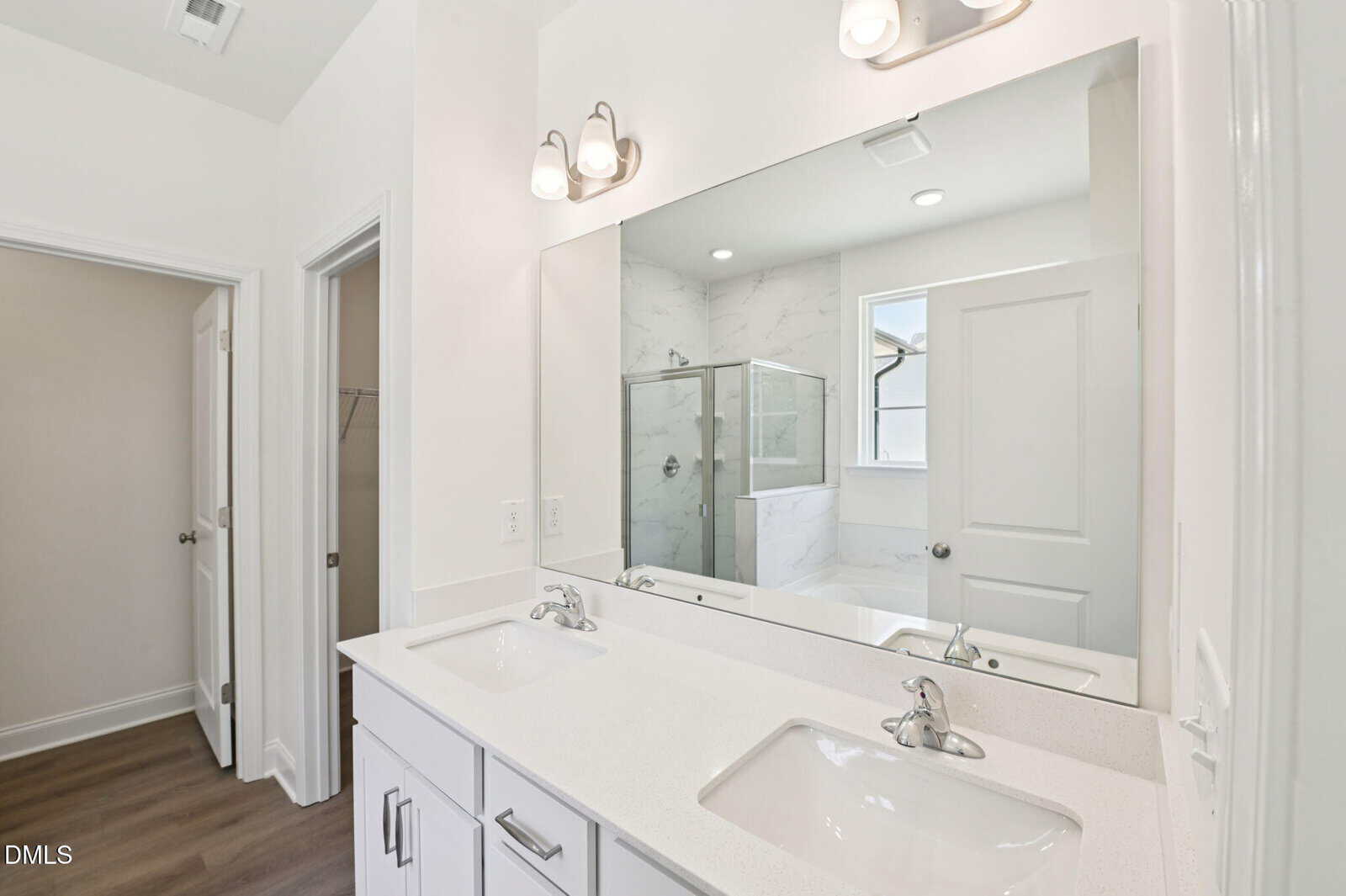 A modern bathroom with a large mirror above a white double-sink vanity, chrome faucets, overhead lights, and a glass shower enclosure in the background. The floor is wood, and walls are white.