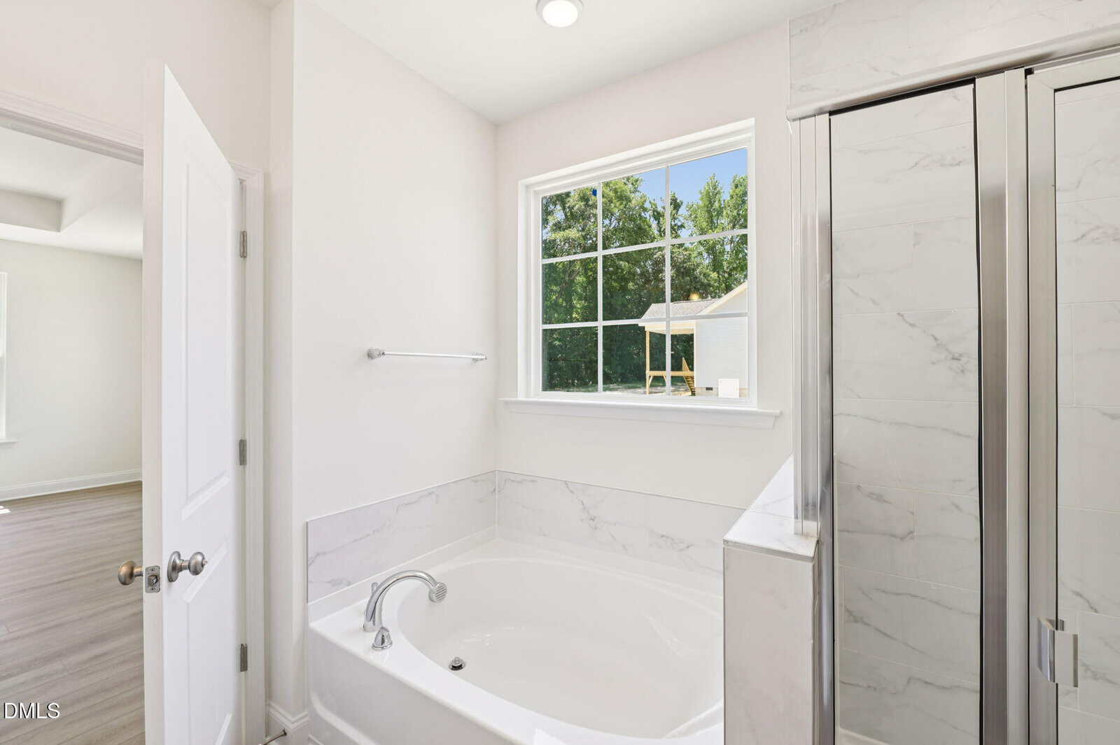 Bright bathroom with a white tub, silver faucet, glass shower, and large window showing greenery outside. Walls and tiles are white with marble accents. Door is open to an adjacent room.