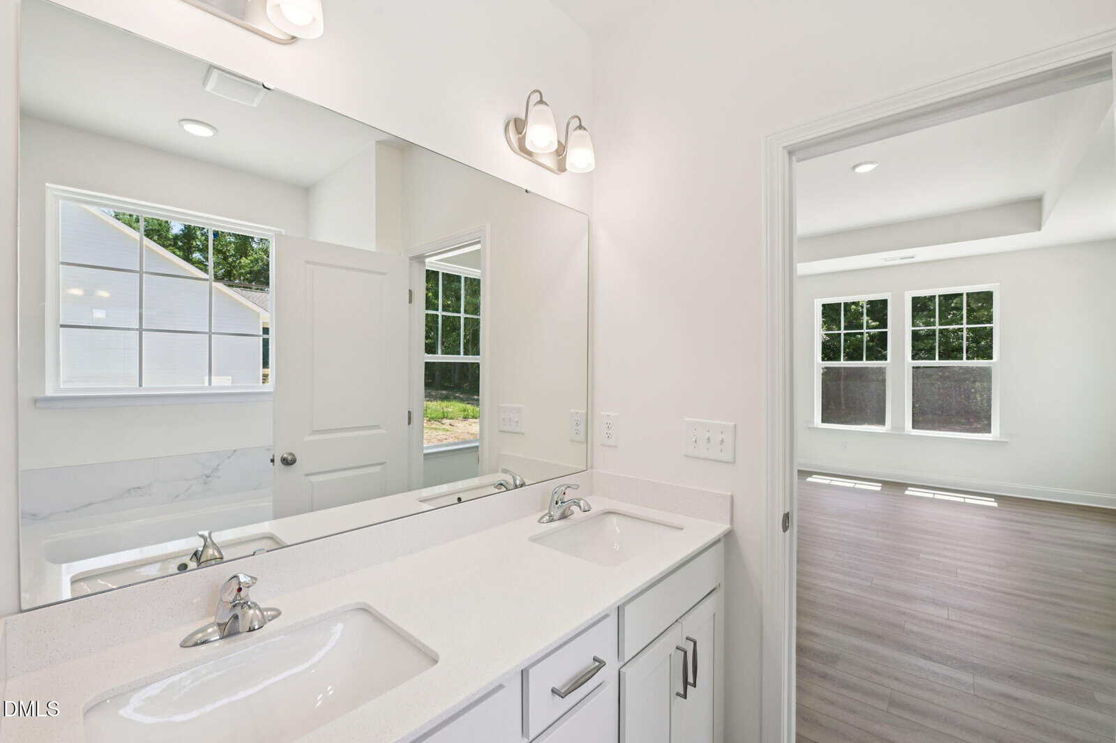 Bright bathroom with double sinks, large mirror, white cabinets, and simple lighting fixtures; door opens to a sunlit room with large windows and wood flooring.