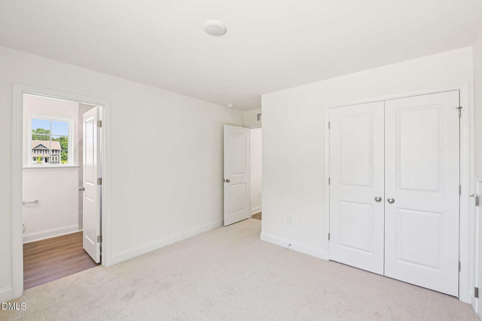 Bright, empty bedroom with white walls, beige carpet, a double-door closet, and an open door leading to a bathroom with a window showing a house and greenery outside.