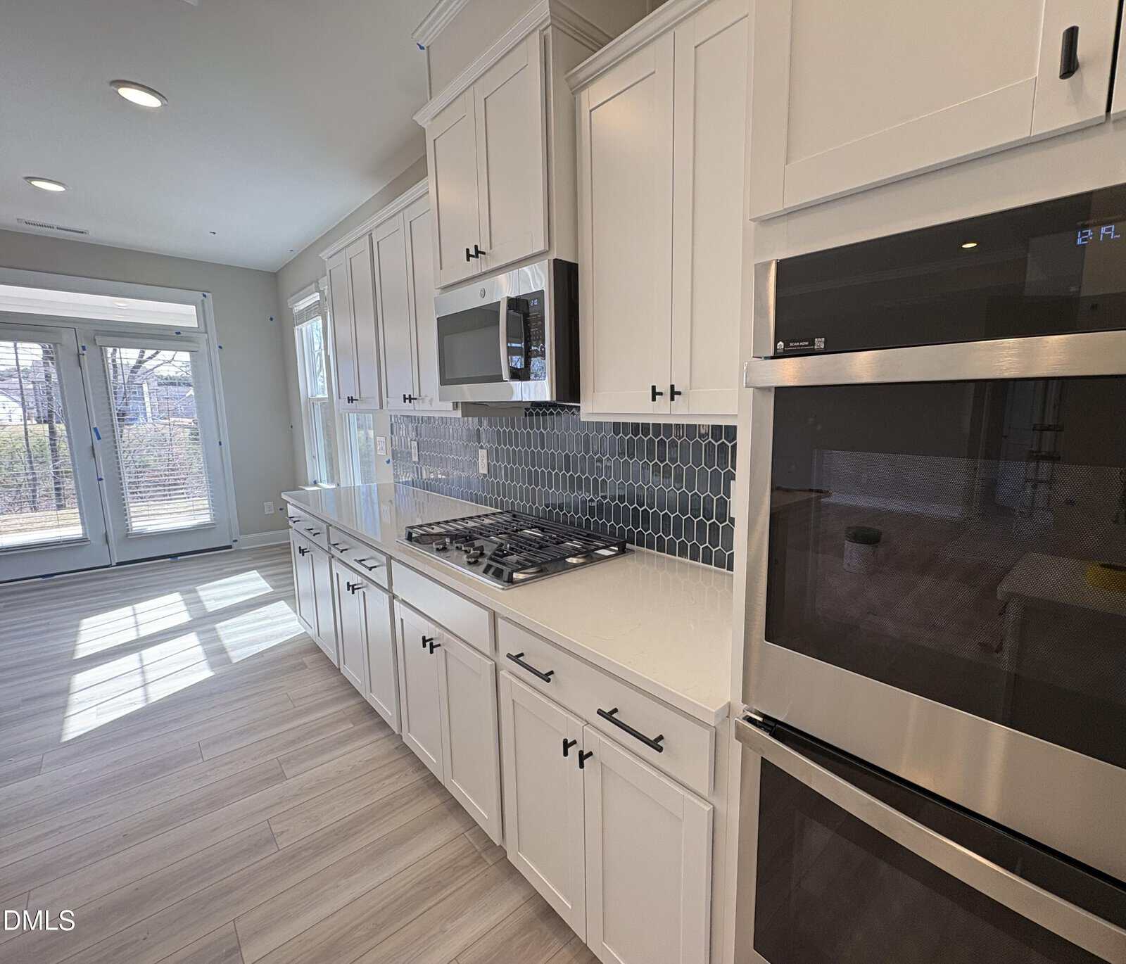 Modern kitchen with white cabinets, stainless steel appliances, a built-in microwave and oven, a gas cooktop, hexagonal tile backsplash, and light wood flooring. Large windows let in natural light.