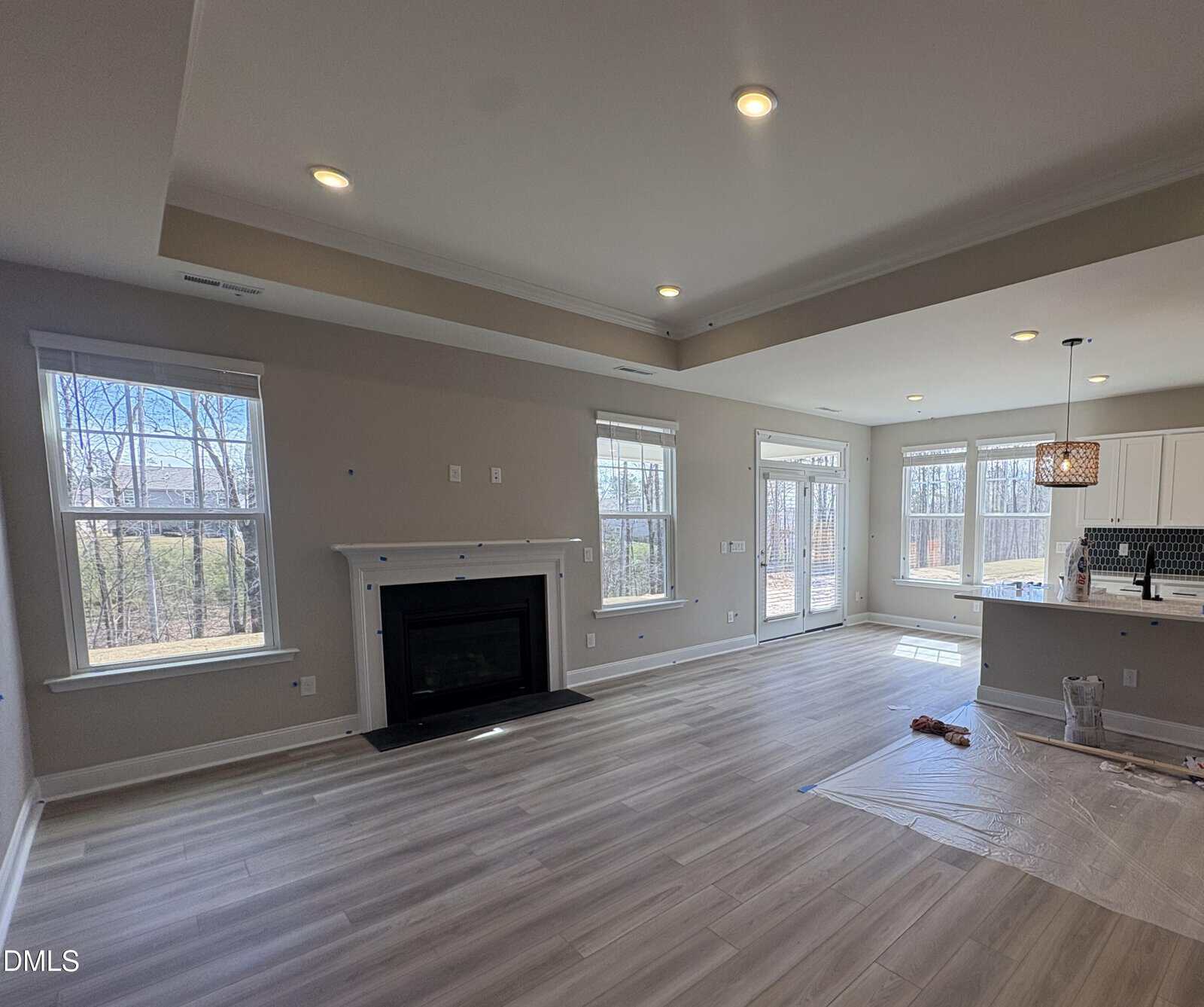 Bright, empty living room with light wood floors, recessed lighting, a black fireplace, and large windows. An adjacent kitchen area and a door to the outside are visible. Some painting supplies are on the floor.