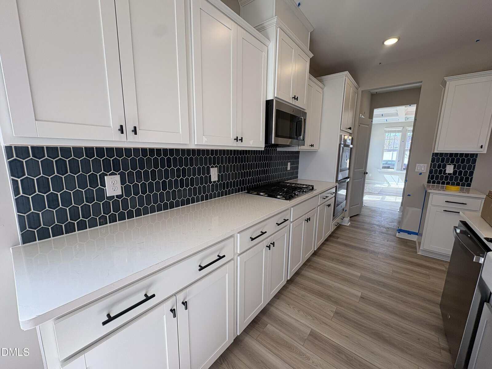 Modern kitchen with white cabinets, black hexagonal tile backsplash, stainless steel appliances, and light wood flooring. The countertop is white and spacious, creating a clean and contemporary look.