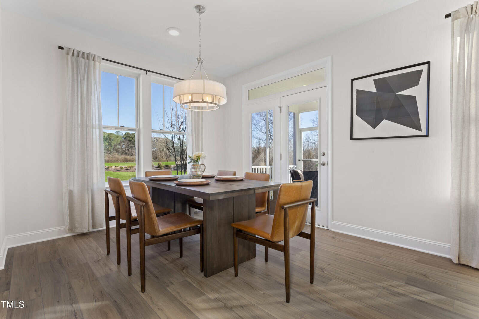 A modern dining room with a dark wood table, six brown leather chairs, a round chandelier, large windows with beige curtains, a door leading outside, and abstract art on the white wall.