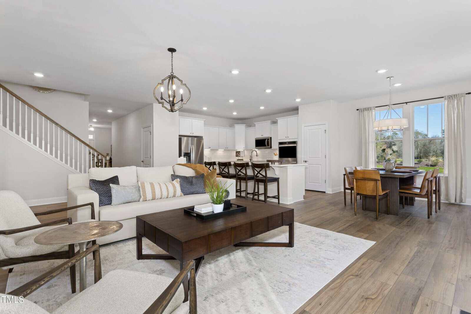 Modern open-concept living and dining area with white walls, wood flooring, a beige sofa, armchairs, dark wood coffee table, kitchen island with barstools, and a dining table by large windows with natural light.
