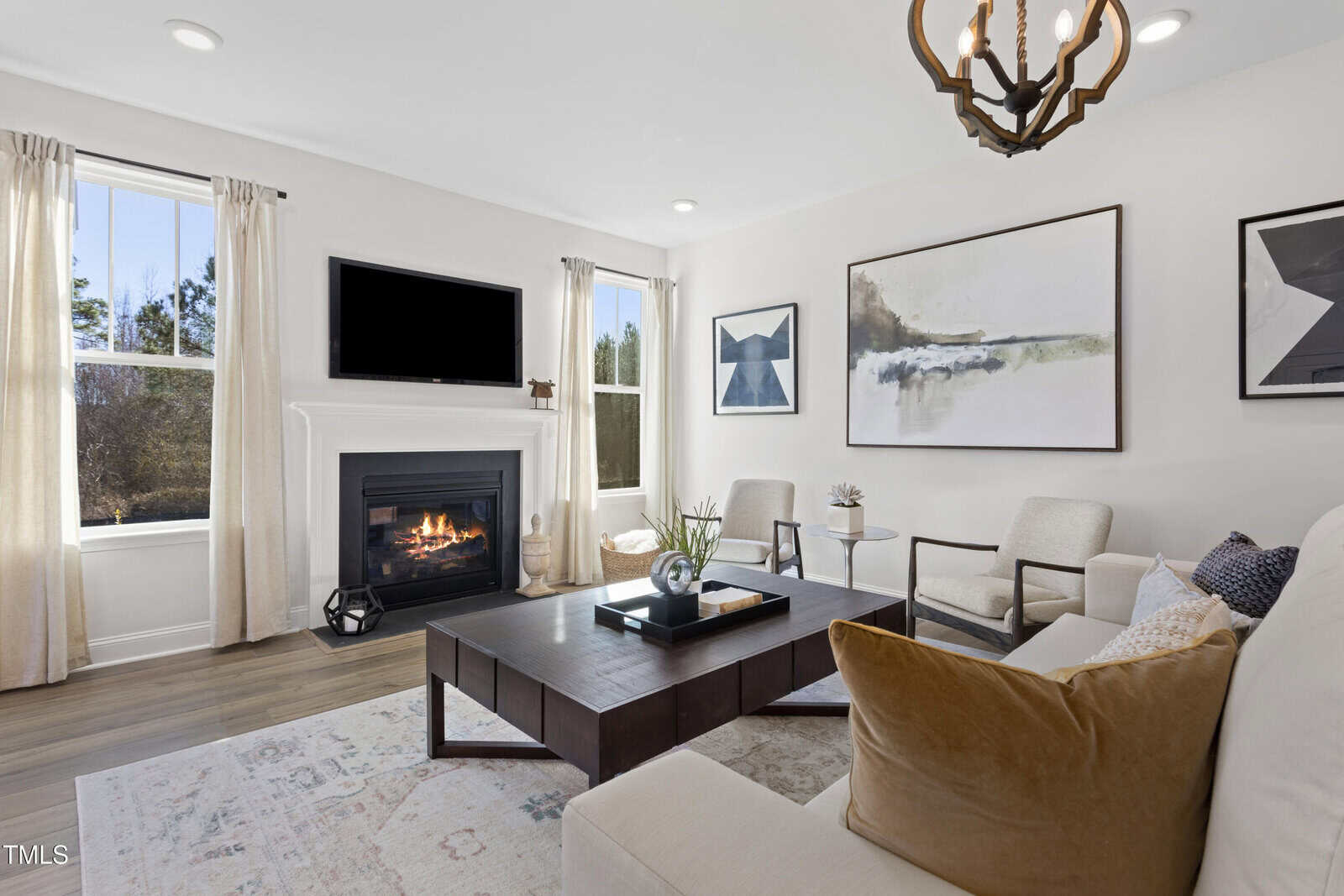 A modern living room with a white sofa, beige armchairs, and a wooden coffee table. A fireplace with a TV above it sits between two windows with light curtains. Abstract art hangs on the white walls.