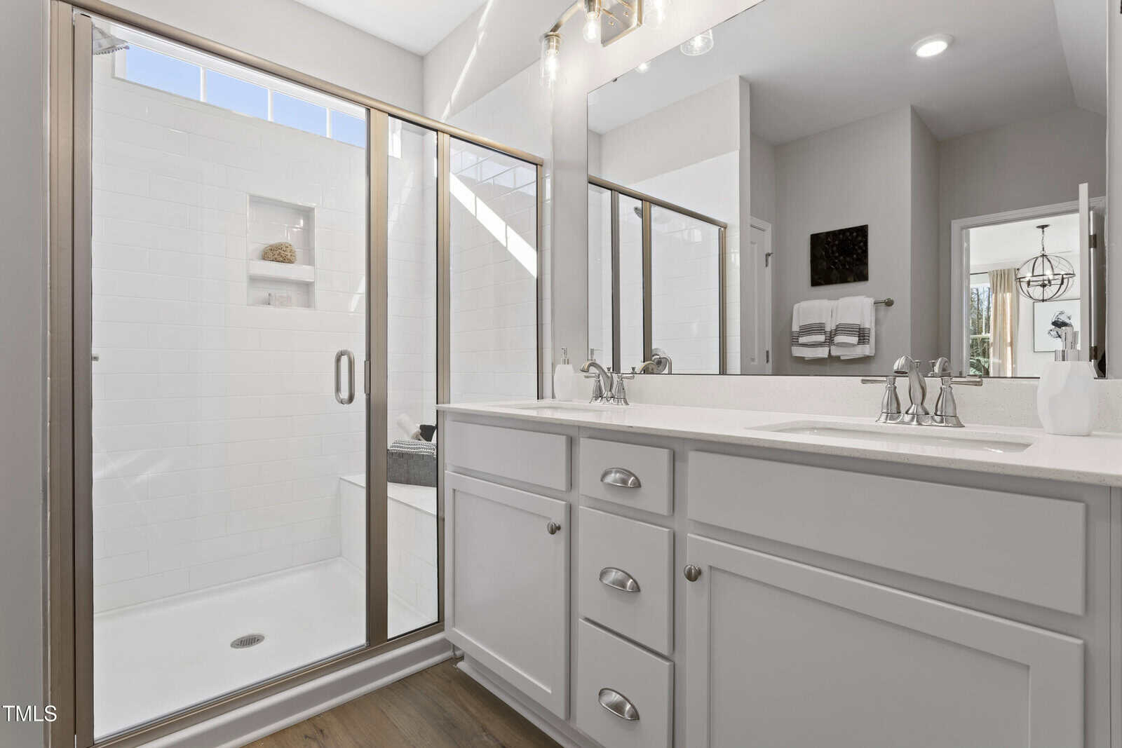 Modern bathroom with a large glass-enclosed shower, white vanity with double sinks and chrome fixtures, white cabinets, wood flooring, and a wall mirror reflecting a bright, clean space.