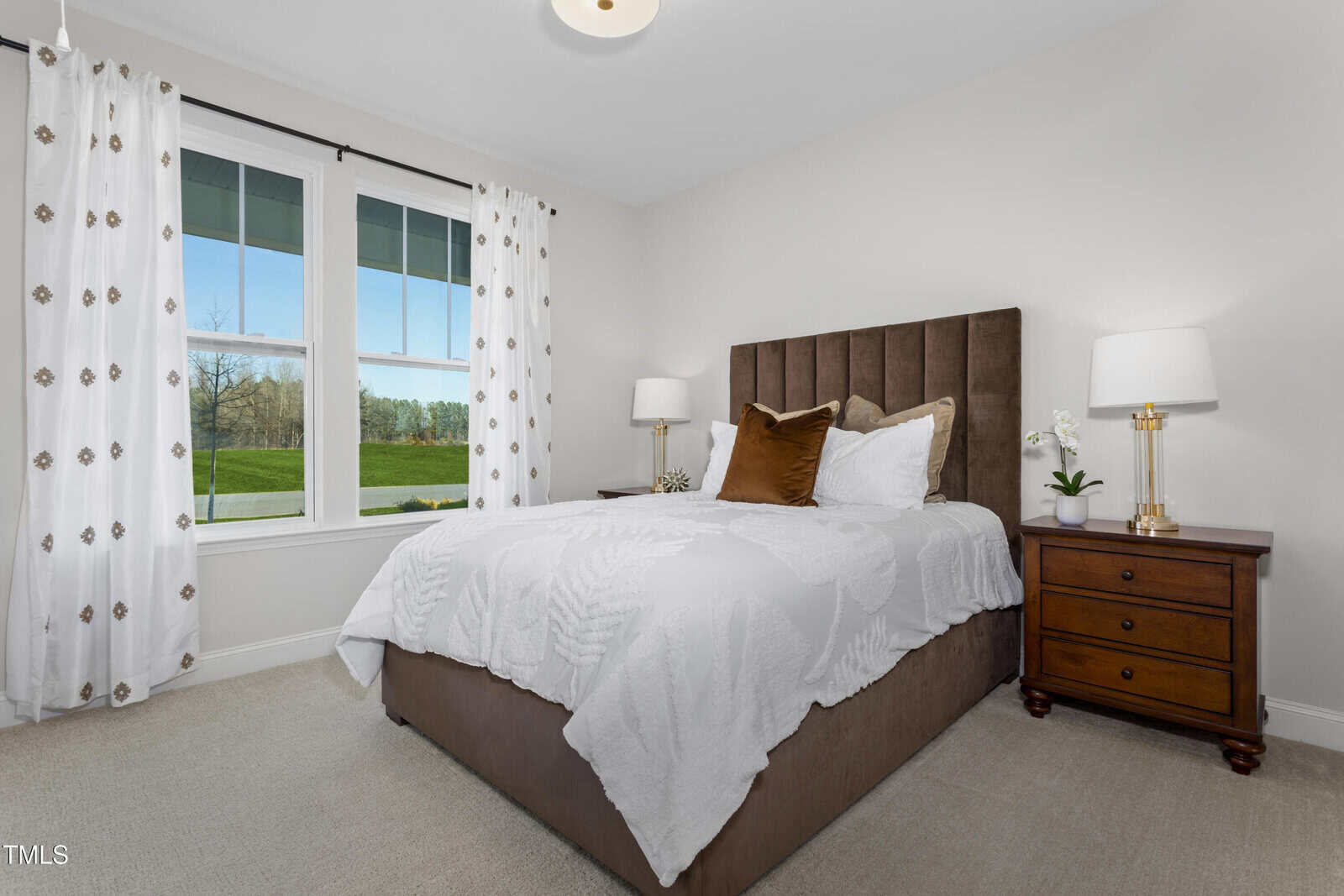 A bright bedroom features a brown upholstered bed with white bedding, a brown accent pillow, two wooden nightstands with lamps, and a window with white curtains overlooking green grass and trees.