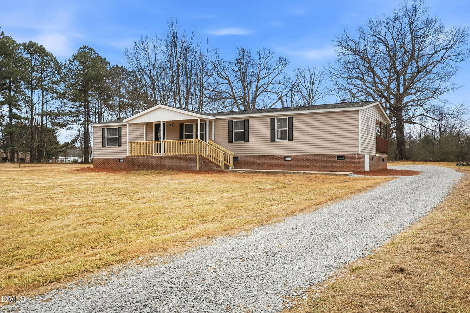 A single-story beige house with black shutters and a brick foundation, featuring a small covered front porch. A gravel driveway curves from the foreground to the right side of the house, with trees and open lawn surrounding it.