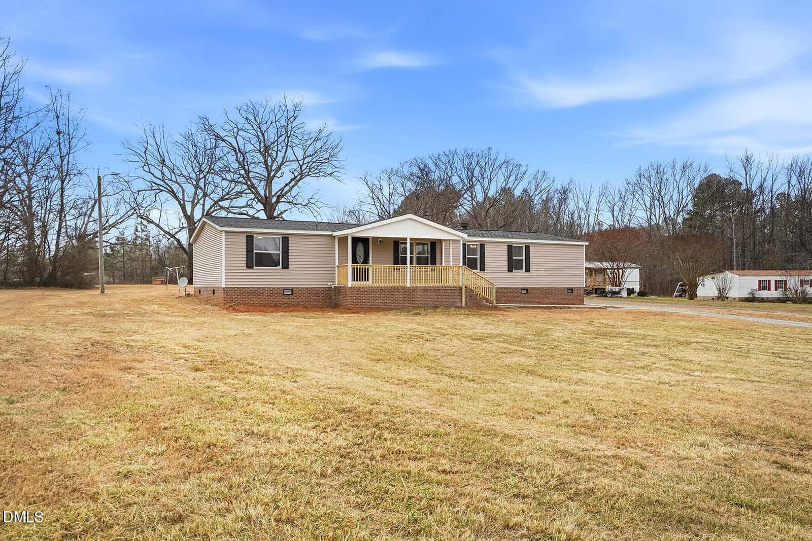 A single-story beige manufactured home with a covered front porch and wooden steps, set on a large grassy yard with trees in the background under a blue sky.