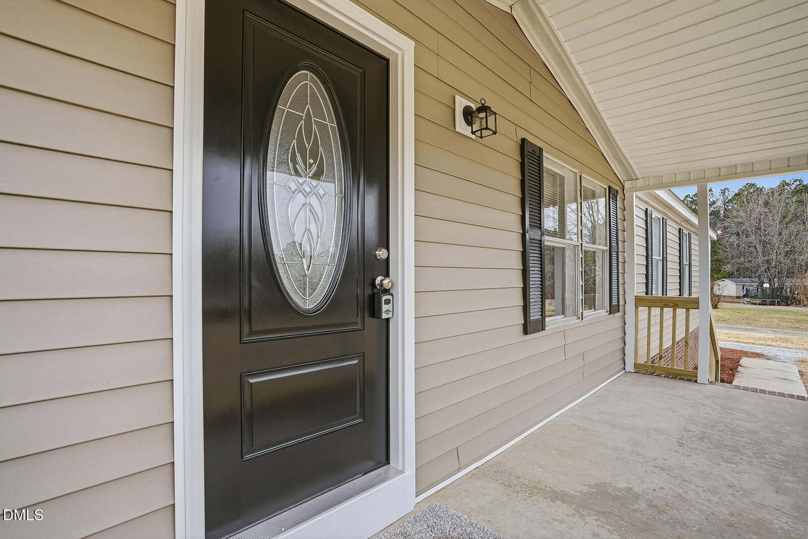 A black front door with an oval glass insert on a tan house with vinyl siding, a covered porch, a wall-mounted light, black shutters, and a concrete walkway. Trees and a road are visible in the background.