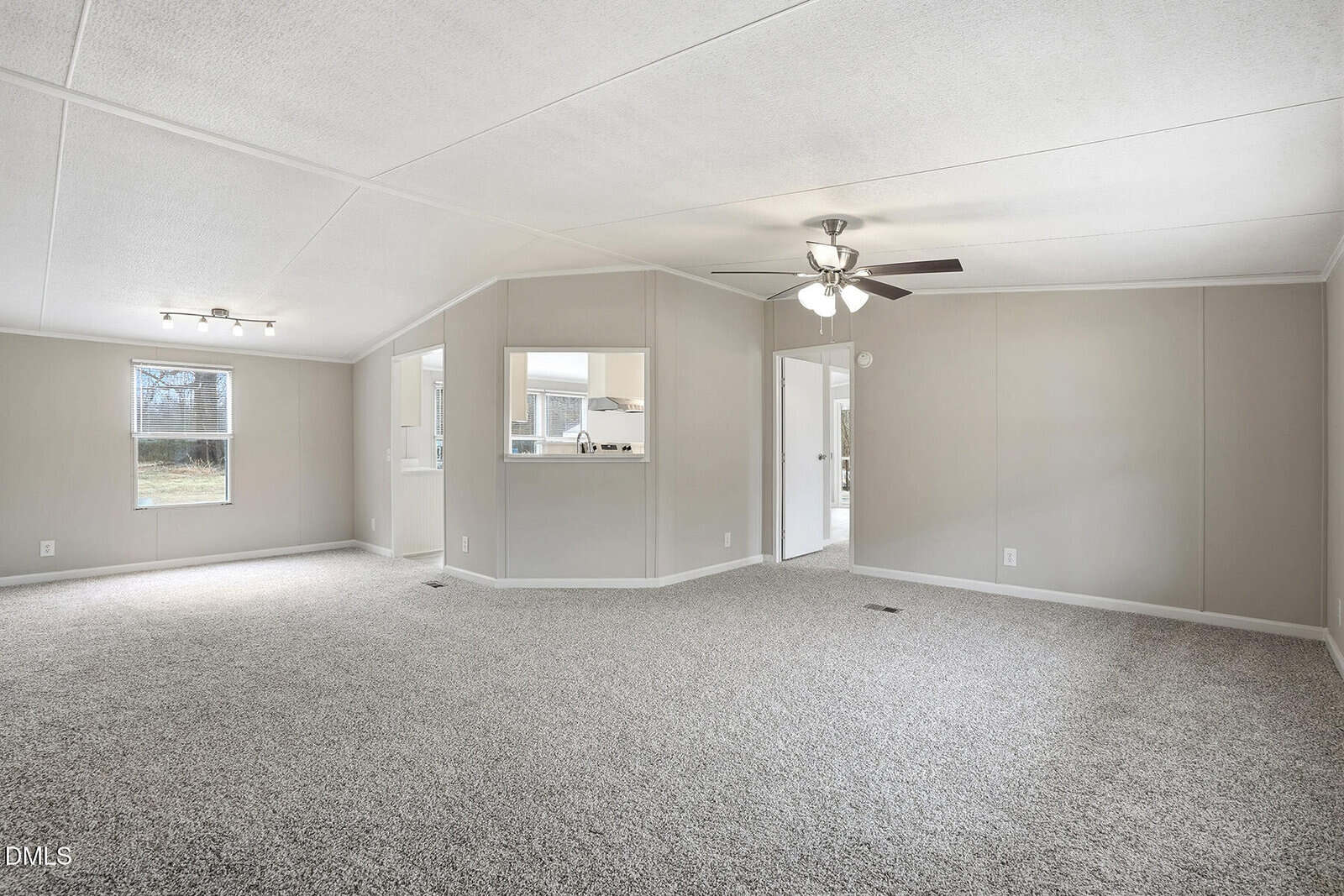 Spacious living room with light gray carpet, beige walls, white ceiling, a ceiling fan, and large windows. There is an open doorway leading to a kitchen and another room, with natural light entering from outside.