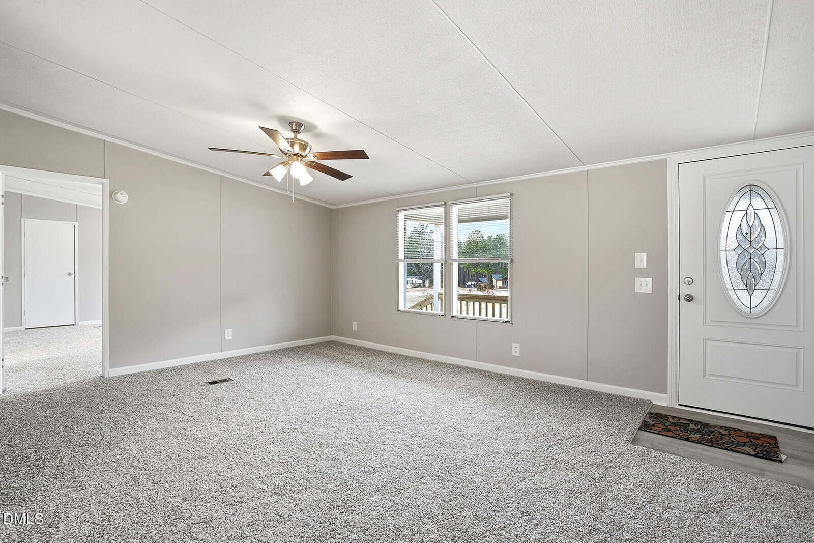 A spacious living room with light gray carpet, beige walls, a ceiling fan with lights, two front windows, and a white door with an oval glass insert. An open doorway leads to another room.